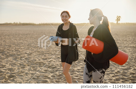 Two smiling fit women walking on the beach with their yoga mats after doing morning gym Two smiling fit women walking on the beach with their yoga mats after doing morning gym 111456182
