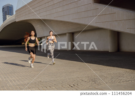 Two active women athlete running side by side along an outdoor track on modern building background Two active women athlete running side by side along an outdoor track on modern building background 111456204