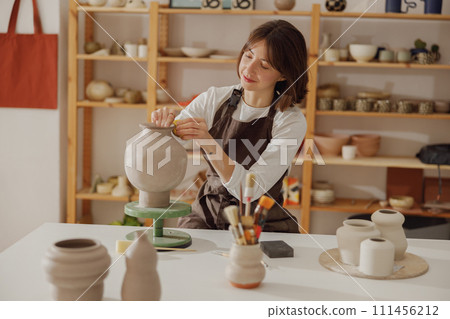 Smiling young female ceramist wearing apron work with unfired clay vase in pottery studio 111456212
