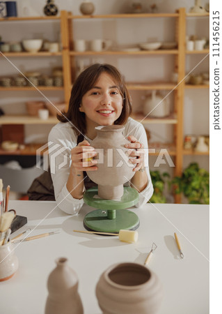 Smiling entrepreneur crafts woman holding mug in pottery studio while looking at camera Smiling entrepreneur crafts woman holding mug in pottery studio while looking at camera 111456215