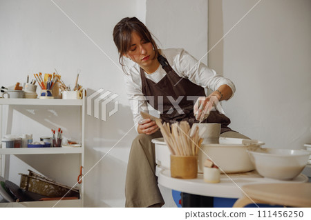 Focused female artisan in apron sitting on bench with pottery wheel and making clay pot 111456250