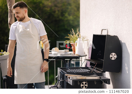 Side view of handsome man try how hot is BBQ grill. Man in apron make dinner outdoor 111467020