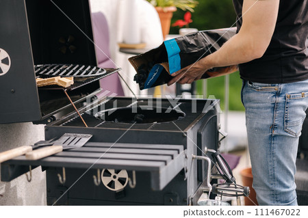 Close-up of man pours out charcoal into BBQ grill on terrace. Male preparing for backyard barbecue Close-up of man pours out charcoal into BBQ grill on terrace. Male preparing for backyard barbecue 111467022