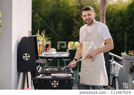 Portrait of handsome young chief in apron stand by the BBQ grill and smile. Male look into camera and rubs hands. Portrait of handsome young chief in apron stand by the BBQ grill and smile. Male look into camera and rubs hands. 111467029