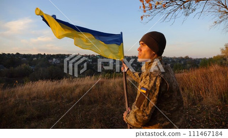 Young man in military uniform waving flag of Ukraine against beautiful sunset at background. Male ukrainian army soldier lifted national banner at countryside. Victory against russian aggression Young man in military uniform waving flag of Ukraine against beautiful sunset at background. Male ukrainian army soldier lifted national banner at countryside. Victory against russian aggression 111467184