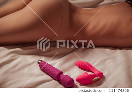 pink sex vibrator and a vacuum stimulator lie next to a sexy woman girl with slim body in panties on white sheet on bed in the bedroom. Sex toys for female satisfaction and orgasm pink sex vibrator and a vacuum stimulator lie next to a sexy woman girl with slim body in panties on white sheet on bed in the bedroom. Sex toys for female satisfaction and orgasm 111474206