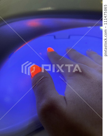 Female hand with neon orange manicure inside a blue UV lamp, drying gel polish. Manicure process in a beauty salon, drying stage. Salon procedure. Female hand with neon orange manicure inside a blue UV lamp, drying gel polish. Manicure process in a beauty salon, drying stage. Salon procedure. 111475865