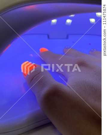 Female hand with neon orange manicure inside a blue UV lamp, drying gel polish. Manicure process in a beauty salon, drying stage. Salon procedure. 111475874