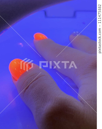 Female hand with neon orange manicure inside a blue UV lamp, drying gel polish. Manicure process in a beauty salon, drying stage. Salon procedure. Female hand with neon orange manicure inside a blue UV lamp, drying gel polish. Manicure process in a beauty salon, drying stage. Salon procedure. 111475882