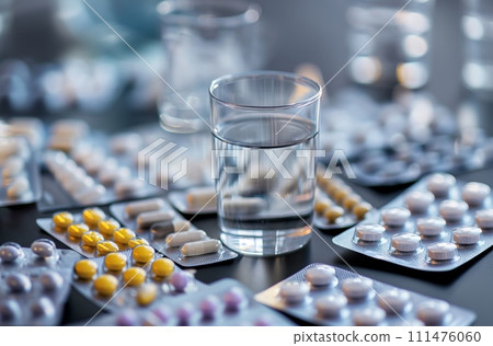 AI-generated content. Several blister packs containing a variety of medical pills and capsules are spread out on a grey tabletop, a glass of water 111476060