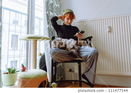 teenage girl sitting on chair, holding cat and looking at phone 111477544