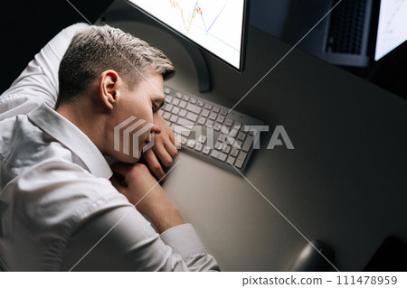 High-angle view of tired male trader asleep on computer desk late at night. Exhausted businessman sleeping with head on keyboard while working overnight on computer. Concept of overwork and deadline 111478959