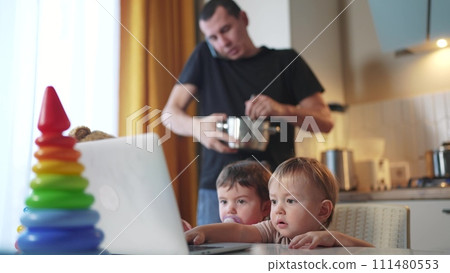 father cooking in the kitchen remote work. baby twins playing with laptop in the kitchen. father working lifestyle at home in the kitchen stirring food in the pan. remote work business concept 111480553