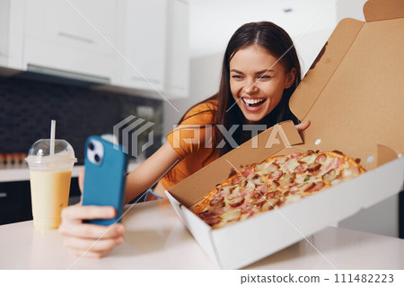 Woman holding pizza taking selfie with phone and pizza box, enjoying delicious Italian meal and capturing the moment on camera 111482223