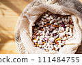 Multi-colored beans spilling out from an open, natural fiber mesh eco bag onto wooden surface with sunlight shadows. Healthy eating in sustainable packaging 111484755