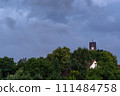 Idyllic village scene with a church tower rising above lush green trees against dramatic overcast sky. Contrast between darkened sky and natural greenery creates a tranquil yet moody atmosphere. 111484758
