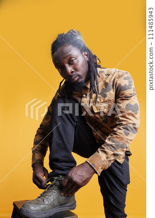 Serious young adult preparing shoes tying shoelace, preparing for long walk. African american man wearing fashionable clothes and sneakers standing in studio posing over yellow background. 111485943