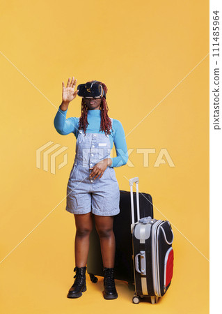 Young adult having fun with vr glasses in studio, posing over orange background with virtual reality headset and trolley bags. Female traveller using modern goggles with 3d vision. 111485964