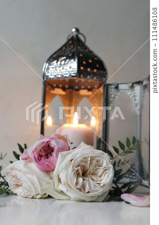 Silver Moroccan, Arabic lantern with burning candle. Pink roses flowers on white table. Festive still life for muslim holiday Ramadan Kareem or boho wedding. Blurred background, vertical. 111486108