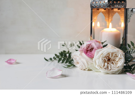 Burning silver Moroccan, Arabic lantern with pink roses flowers and green leaves on white table. Festive still life for muslim holiday Ramadan Kareem or boho wedding. Blurred background. 111486109