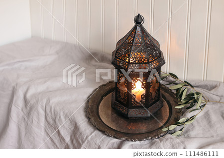 Burning black Moroccan, Arabic lantern with silver tray, olive tree twigs on table with linen tablecloth. White wooden wall background. Muslim holiday Ramadan Kareem home still life, iftar dinner. Burning black Moroccan, Arabic lantern with silver tray, olive tree twigs on table with linen tablecloth. White wooden wall background. Muslim holiday Ramadan Kareem home still life, iftar dinner. 111486111