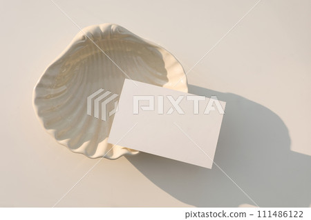 Summer neutral wedding stationery. Blank greeting, RSVP or business card mockup in sunlight. Invitation, template. Beige sea shell plate. White table background. Elegant composition. Flatlay, top view Summer neutral wedding stationery. Blank greeting, RSVP or business card mockup in sunlight. Invitation, template. Beige sea shell plate. White table background. Elegant composition. Flatlay, top view 111486122
