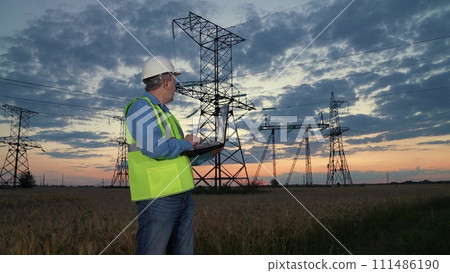 Technician setups network with laptop standing against power transmission lines in sunset field. Mature engineer reports about electrical system via laptop at power distribution substation in evening Technician setups network with laptop standing against power transmission lines in sunset field. Mature engineer reports about electrical system via laptop at power distribution substation in evening 111486190