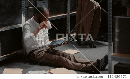 Policeman sitting on floor searching for clues in archived case files, working in police incident room. Professional officer conducting criminal investigation and doing suspect background check. 111486371