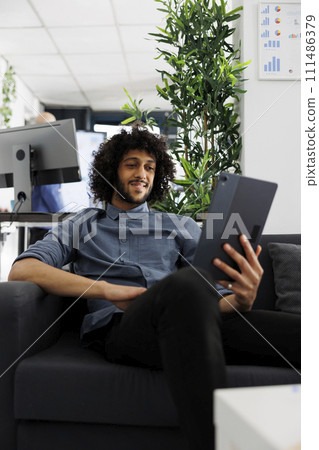 Arab business employee telecommuting, chatting in online meeting using digital tablet. Smiling relaxed start up entrepreneur videconferencing while sitting on couch in office 111486379