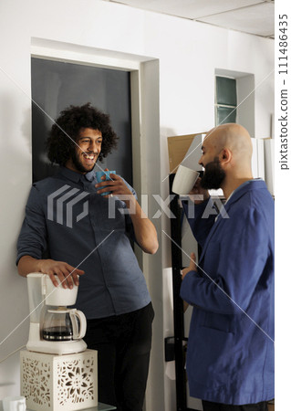 Two smiling arab businessmen having coffee break and talking in business office. Company employees standing with tea mugs while laughing and chatting in coworking space workplace Two smiling arab businessmen having coffee break and talking in business office. Company employees standing with tea mugs while laughing and chatting in coworking space workplace 111486435