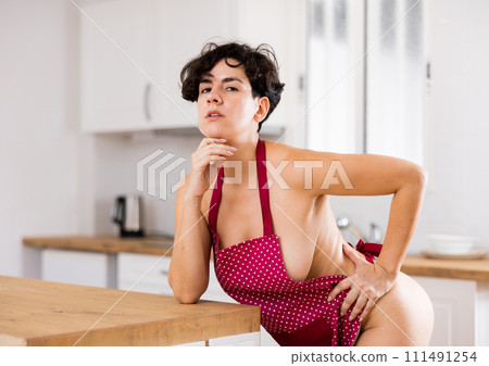 Portrait of flirting female in red apron posing playfully, leaning on countertop in kitchen 111491254