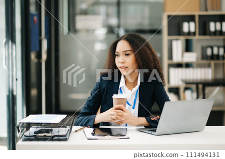 Confident beautiful businesswoman typing laptop and digital tablet while holding coffee . 111494131