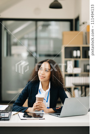 Confident beautiful businesswoman typing laptop and digital tablet while holding coffee . 111494133
