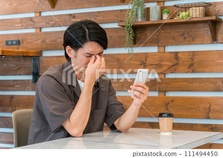 一名年輕的亞洲男子在咖啡館、家裡等使用智慧型手機(眼睛疲勞、眼睛疲勞、眼睛乾澀、疲勞) 一名年輕的亞洲男子在咖啡館、家裡等使用智慧型手機(眼睛疲勞、眼睛疲勞、眼睛乾澀、疲勞) 111494450