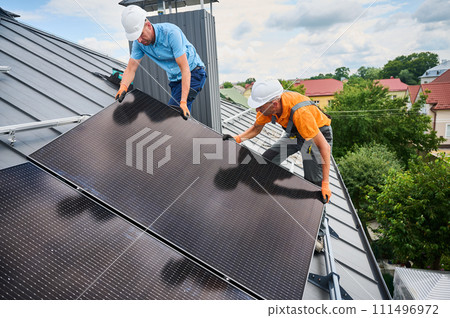 Workers building solar panel system on rooftop of house. Two men installers in helmets installing photovoltaic solar module outdoors. Alternative, green and renewable energy generation concept. Workers building solar panel system on rooftop of house. Two men installers in helmets installing photovoltaic solar module outdoors. Alternative, green and renewable energy generation concept. 111496972