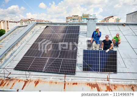 Workers building solar panel system on metal rooftop of house. Three men installers installing photovoltaic solar module outdoors. Alternative, green and renewable energy generation concept. Workers building solar panel system on metal rooftop of house. Three men installers installing photovoltaic solar module outdoors. Alternative, green and renewable energy generation concept. 111496974