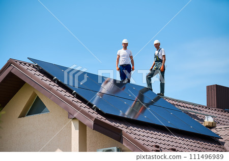 Workers building solar panel system on metal rooftop of house. Portrait of engineers standing on rooftop while installing photovoltaic solar module outdoors. Renewable energy generation concept. 111496989