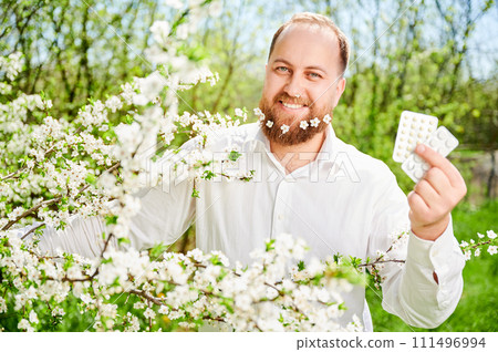 Man allergic suffering from seasonal allergy at spring. Smiling man with small white flowers dotted in beard, holding pack of pills, posing in blossoming garden. Antihistamine medication concept 111496994