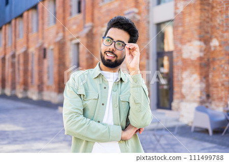 Interested indian man with beard touching eyeglasses and looking aside while passing brick building. Carefree young person having walk in old district while doing sightseeing and getting impressed. Interested indian man with beard touching eyeglasses and looking aside while passing brick building. Carefree young person having walk in old district while doing sightseeing and getting impressed. 111499788