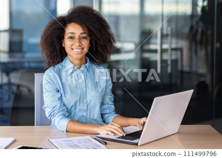 Cheerful diverse woman smiling at camera while keeping hands on portable computer keyboard in workplace. Remote digital designer feeling satisfied with completing massive high paying task online. Cheerful diverse woman smiling at camera while keeping hands on portable computer keyboard in workplace. Remote digital designer feeling satisfied with completing massive high paying task online. 111499796
