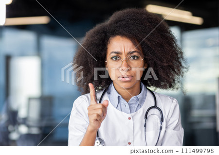 Serious african american doctor in white lab coat raising index finger with rigorous facial expression. Experienced cardiologist emphasising harm of smoking to cardiovascular system for adults. Serious african american doctor in white lab coat raising index finger with rigorous facial expression. Experienced cardiologist emphasising harm of smoking to cardiovascular system for adults. 111499798