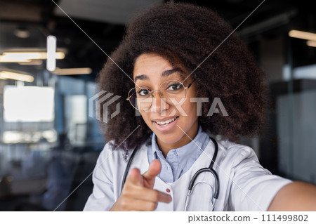 Diverse medic student in eyeglasses with bushy hairdo pointing at camera on blurred background. Pretty female intern recording video about importance of regular body checks for disease preventing. 111499802