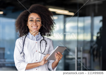 Responsible hispanic nurse with curly hair and stethoscope over neck smiling friendly and holding digital tablet. Optimistic hospital worker checking online registration of clients for procedures. 111499806