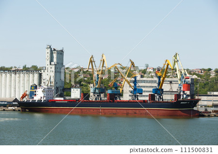 Cargo ship docked at industrial port. Maritime vessel loading, unloading freight containers at harbor, daytime commercial distribution hub. Bulk carrier, cranes against clear blue sky, logistics. 111500831
