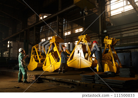 Industrial workers inspect yellow grapples in factory setting. Heavy equipment manufacturing process, teamwork with safety helmets. Quality control in metal fabrication, engineered machinery parts. 111500833
