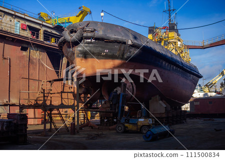 Industrial workers repair fishing boat in dry dock. Maintenance of marine vessel hull, welding, painting under blue sky. Shipyard activity, nautical transport services. 111500834