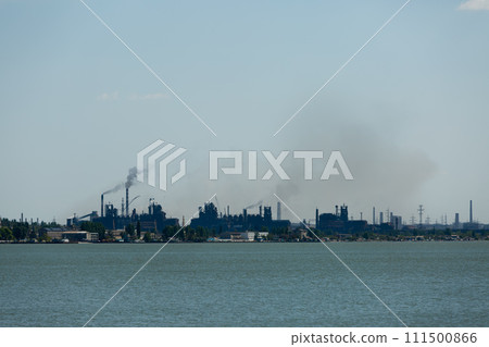 Industrial skyline dominated by metallurgical plant emissions, smoke billowing against sky, symbolizing eco pollution issues, with water foreground. 111500866
