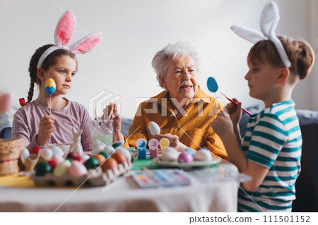 Grandmother with little kids decorating easter eggs at home. Tradition of painting eggs with brush and easter egg dye. Grandmother with little kids decorating easter eggs at home. Tradition of painting eggs with brush and easter egg dye. 111501152