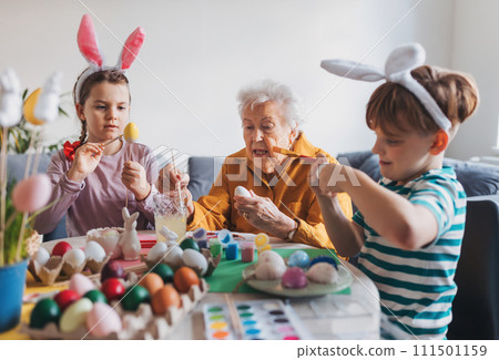 Grandmother with little kids decorating easter eggs at home. Tradition of painting eggs with brush and easter egg dye. 111501159