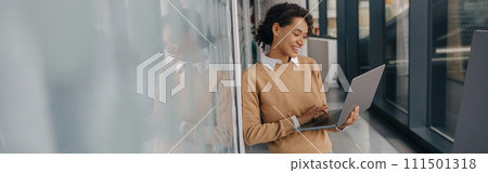 Stylish business woman using laptop while standing on modern office background Stylish business woman using laptop while standing on modern office background 111501318